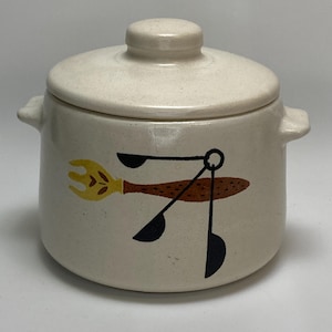 May include: A cream-colored ceramic jar with a lid, featuring a whimsical design of kitchen utensils in brown, yellow, and black. The jar has two small handles and a rounded shape, perfect for kitchen storage.