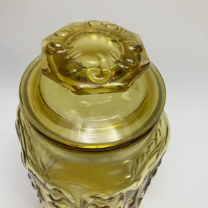 May include: A vintage amber glass jar with a lid. The jar has a decorative, embossed pattern on the sides and lid. The lid is hexagonal with a floral design. The jar is made of transparent glass.