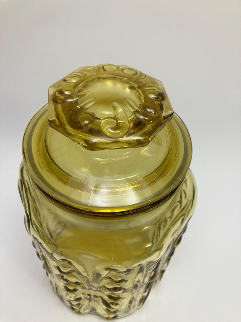 May include: A vintage amber glass jar with a lid. The jar has a decorative, embossed pattern on the sides and lid. The lid is hexagonal with a floral design. The jar is made of transparent glass.