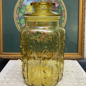 May include: A vintage yellow glass jar with a lid. The jar has a square shape with embossed floral designs. The lid is round and fits snugly on top. The jar is sitting on a white lace doily. A framed picture is in the background.