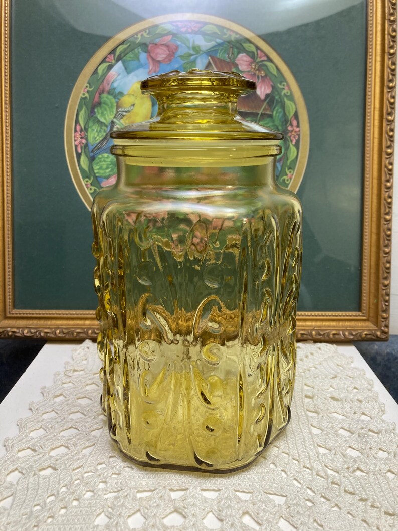 May include: A vintage yellow glass jar with a lid. The jar has a square shape with embossed floral designs. The lid is round and fits snugly on top. The jar is sitting on a white lace doily. A framed picture is in the background.