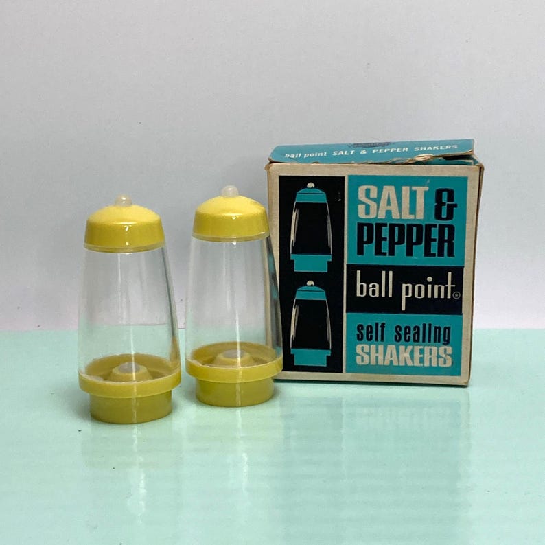 May include: Two clear plastic salt and pepper shakers with yellow lids and bases. The shakers are next to a box that reads "SALT & PEPPER ball point self sealing SHAKERS" in teal and black.