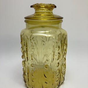 May include: A vintage amber glass jar with a lid. The jar has a rectangular shape with embossed floral and swirl designs. The lid has a decorative knob. The jar is made of transparent glass.