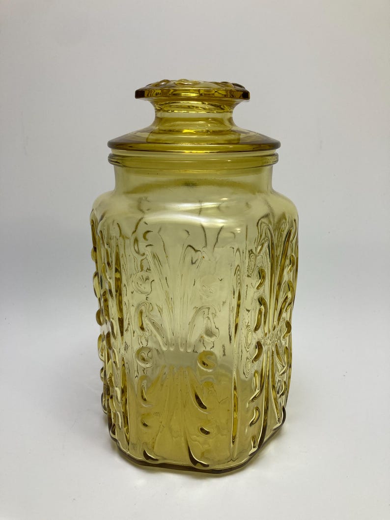 May include: A vintage amber glass jar with a lid. The jar has a rectangular shape with embossed floral and swirl designs. The lid has a decorative knob. The jar is made of transparent glass.