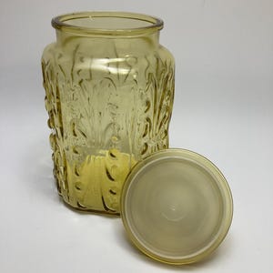 May include: A yellow-tinted glass jar with a matching lid. The jar has a rectangular shape with a wide opening and a decorative, embossed floral pattern. The lid is round and sits next to the jar. The jar is likely for storage.