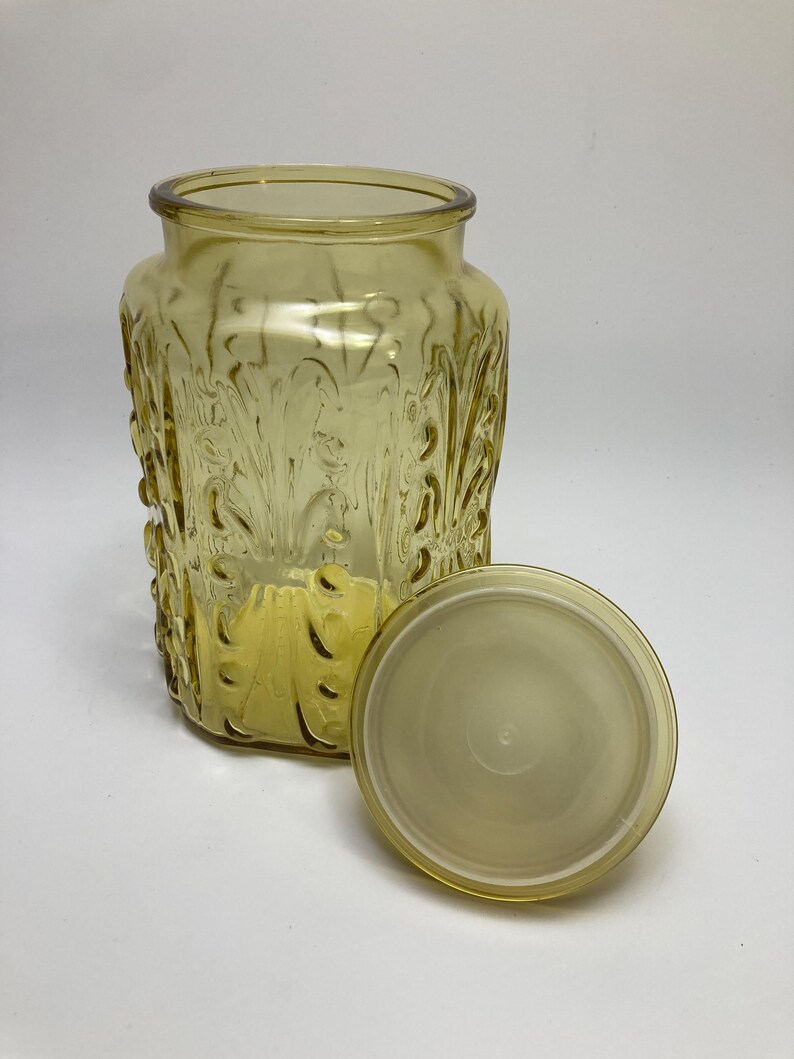 May include: A yellow-tinted glass jar with a matching lid. The jar has a rectangular shape with a wide opening and a decorative, embossed floral pattern. The lid is round and sits next to the jar. The jar is likely for storage.