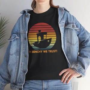 May include: Black t-shirt featuring a boat design inside a sunset circle with orange, yellow, and green stripes. The text "IN BENCHY WE TRUST!" is below the graphic. The shirt is worn with a blue denim jacket and jeans.