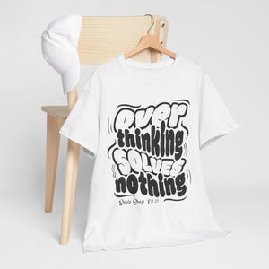 Puede incluir: Camiseta blanca con el texto "Overthinking Solves Nothing" en negrita, negro, con fuente de burbujas. La camiseta está colgada en una percha de madera. Una gorra de béisbol blanca descansa sobre una silla de madera.