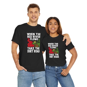 May include: Two people wearing black t-shirts. The shirts feature the text "WHEN THE RED RIVER FLOWS TAKE THE DIRT ROAD" in white, with a red river, yellow sun, and green trees graphic.