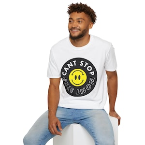 May include: White t-shirt with a black and yellow smiley face graphic. The graphic features the text "CANT STOP WONT STOP" around the smiley face. The shirt is paired with blue jeans.
