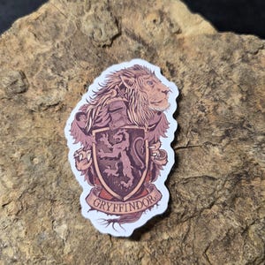 May include: A sticker featuring the Gryffindor house crest from the Harry Potter series. The crest is a shield with a lion, in shades of brown and red, with the word "Gryffindor" on a banner below. The sticker is on a textured, brown surface.