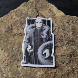 May include: A sticker featuring a black and white illustration of a bald character with a snake wrapped around their neck. The character is wearing a dark robe and holding a small object. The background is a dark, vertical striped design.