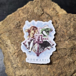 May include: A sticker featuring the Hogwarts crest from the Harry Potter series. The crest is a shield shape with a lion, snake, eagle, and badger, with the word "HOGWARTS" at the bottom.
