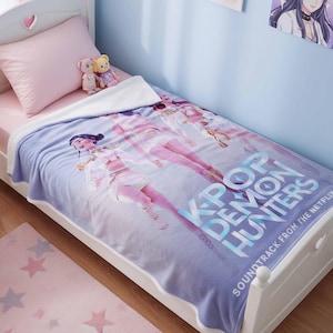 Pode incluir: Uma cama de solteiro branca com lençol e travesseiro rosa, coberta por um cobertor azul e branco com o texto "K-Pop Demon Hunters". O cobertor tem ilustrações de personagens. Um tapete rosa com estampa de estrelas está no chão.