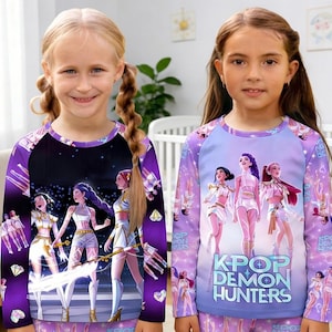 May include: Two young girls wearing matching purple long-sleeved pajama tops and bottoms. The tops feature a graphic of anime-style characters. One shirt says "K-Pop Demon Hunters". The pajamas are a fun and colorful choice for kids.
