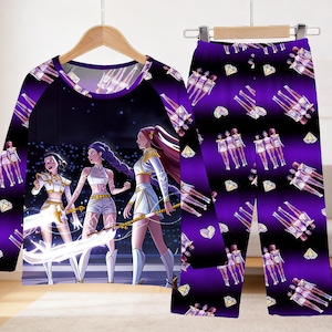 May include: A purple pajama set featuring a long-sleeved top and matching pants. The top has a graphic of three figures in white and gold outfits on a stage. The pants have a repeating pattern of figures, diamonds, and hearts.