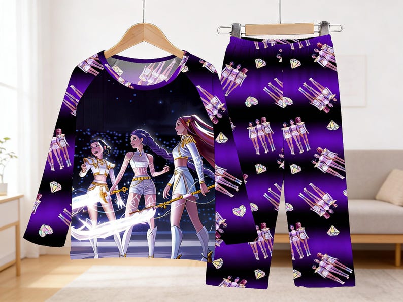 May include: A purple pajama set featuring a long-sleeved top and matching pants. The top has a graphic of three figures in white and gold outfits on a stage. The pants have a repeating pattern of figures, diamonds, and hearts.