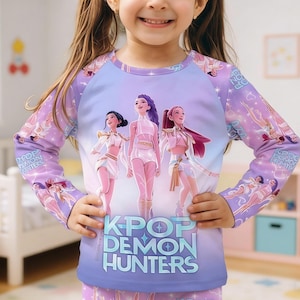 May include: A long-sleeved purple shirt with a graphic of three animated figures. The shirt features the text "K-POP DEMON HUNTERS" in a blue and white font. The sleeves have a repeating pattern of the animated figures. The shirt is worn by a child.