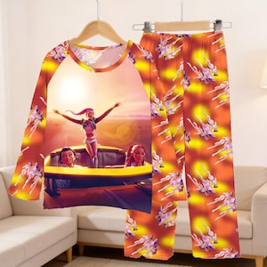 May include: A two-piece pajama set featuring a long-sleeved top and pants. The top has a graphic of a woman with her arms raised in a car, with a sunset background. The pants and sleeves have a repeating pattern of a woman in a car.