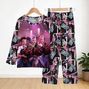 May include: A pajama set featuring a long-sleeved top and pants. The top has a central graphic of three women holding bouquets, with the text "KPOP DEMON HUNTERS". The pants and sleeves have a repeating pattern with the same text and figures.