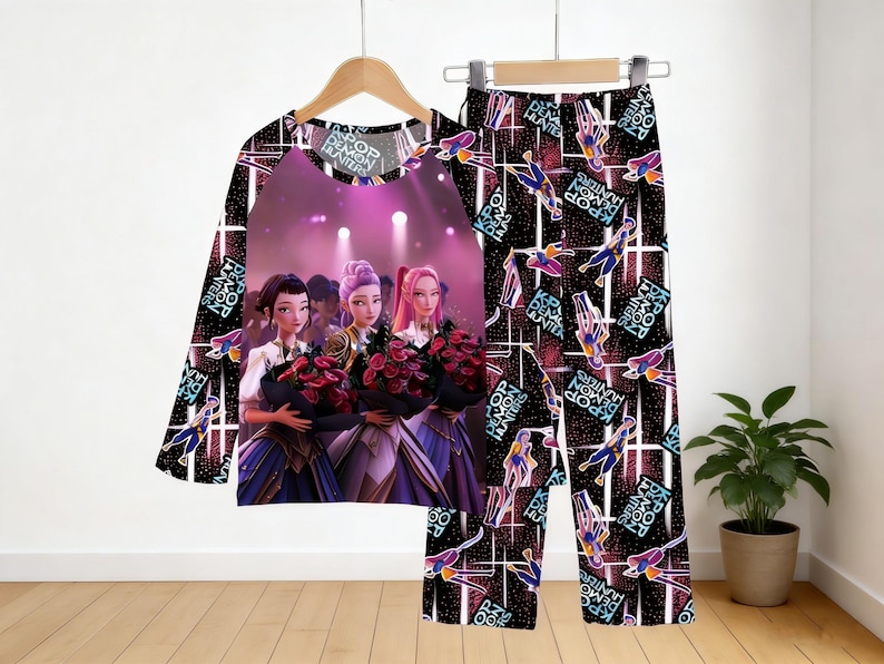 May include: A pajama set featuring a long-sleeved top and pants. The top has a central graphic of three women holding bouquets, with the text "KPOP DEMON HUNTERS". The pants and sleeves have a repeating pattern with the same text and figures.