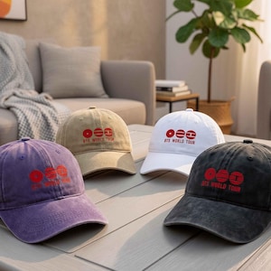 Könnte beinhalten: Vier Baseballkappen in Lila, Beige, Weiß und Schwarz, jede mit einem roten Grafikdesign und dem Text "BTS WORLD TOUR". Die Kappen sind auf einem hellen Tisch angeordnet, der ihre verschiedenen Farben und Designs zeigt.
