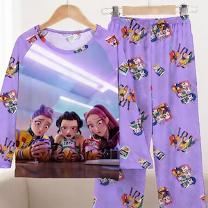 May include: A lavender pajama set featuring a long-sleeved top and matching pants. The top has a large graphic of three animated characters holding snacks. The pants and sleeves are covered in small cartoon illustrations.