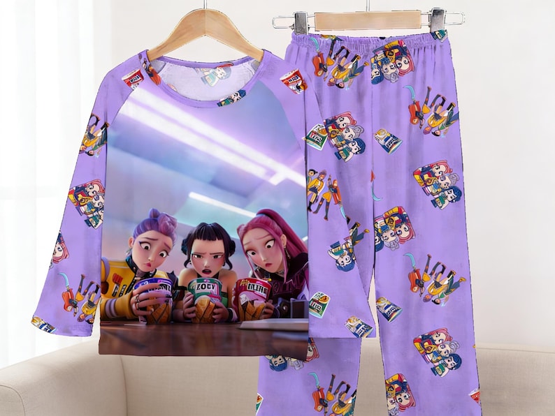 May include: A lavender pajama set featuring a long-sleeved top and matching pants. The top has a large graphic of three animated characters holding snacks. The pants and sleeves are covered in small cartoon illustrations.
