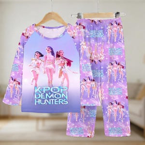 May include: A lilac pajama set featuring a long-sleeved top and pants. The top has a graphic of three anime-style figures with the text "KPOP DEMON HUNTERS." The pants have a repeating pattern of the same figures.