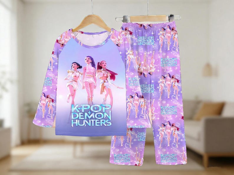 May include: A lilac pajama set featuring a long-sleeved top and pants. The top has a graphic of three anime-style figures with the text "KPOP DEMON HUNTERS." The pants have a repeating pattern of the same figures.