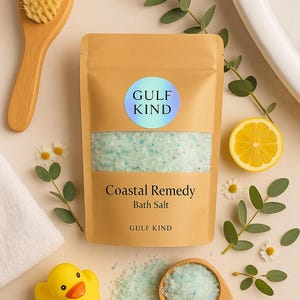 Peut inclure: Un sachet beige de "Coastal Remedy Bath Salt" avec une étiquette bleue et blanche indiquant "Gulf Kind". L'image comprend une brosse en bois, un canard en caoutchouc jaune, une tranche de citron et des brins de verdure.
