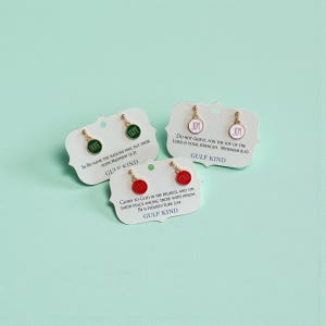 May include: Three pairs of earrings on white cards with scalloped edges. Each earring has a gold-colored post and a circular charm with the words "HOPE", "JOY", or "PEACE" in white. The cards have text from the Bible.