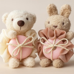 May include: Two plush animal figures: a teddy bear and a bunny. Each holds a pink, heart-shaped soap wrapped in a decorative mesh and tied with twine. The bear is cream-colored, and the bunny is tan.