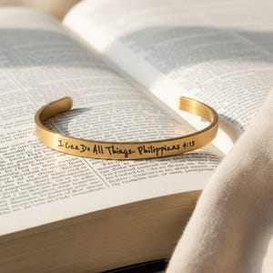 May include: A gold-colored cuff bracelet with the inscription "I Can Do All Things - Philippians 4:13" is displayed open on a book. The bracelet is curved and has a smooth, polished finish. The book's pages are open, with text visible.