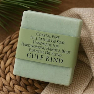 May include: A pale green soap bar with a green label. The label reads "COASTAL PINE" and "GULF KIND." The soap is handmade for hands and body with an essential oil blend. The soap is on a woven mat and a beige fabric.