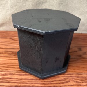 May include: A dark gray, octagonal, stone-like pedestal with a matching top and base. The pedestal has a textured surface and is set on a wooden surface.