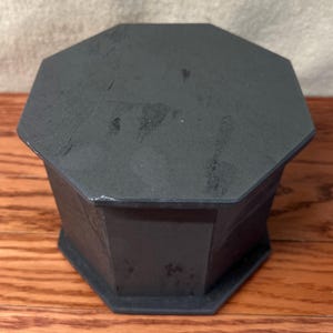 May include: A dark gray, octagonal box with a matching lid. The box is made of a solid material and sits on a wooden surface. The box has a geometric design and a matte finish.