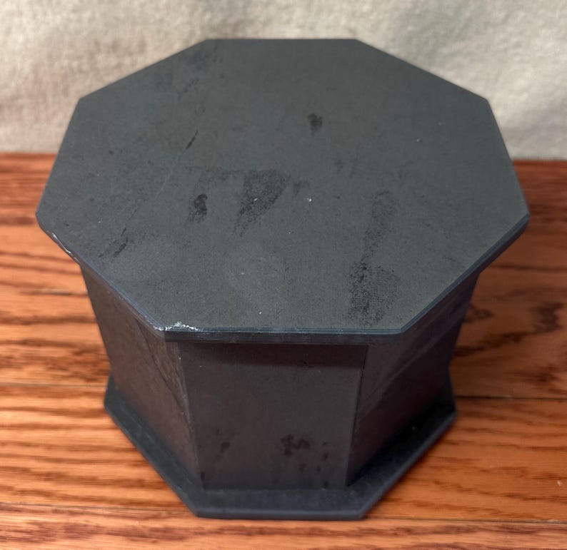 May include: A dark gray, octagonal box with a matching lid. The box is made of a solid material and sits on a wooden surface. The box has a geometric design and a matte finish.
