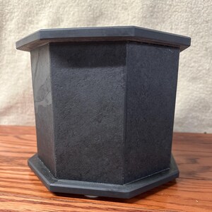 May include: A dark gray, octagonal urn with a textured surface. The urn has a matching octagonal base and a slightly overhanging top. The product is made of a solid, possibly stone-like material.