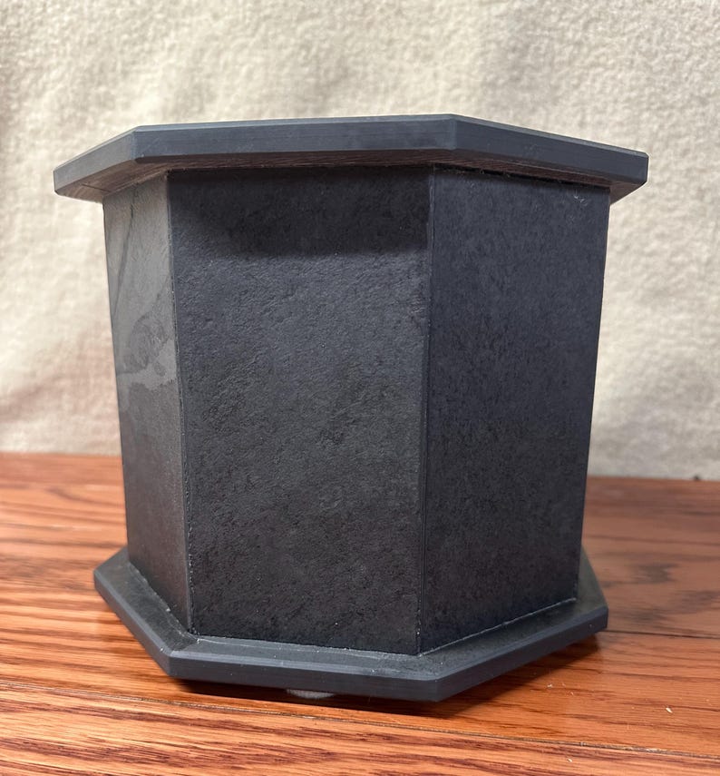 May include: A dark gray, octagonal urn with a textured surface. The urn has a matching octagonal base and a slightly overhanging top. The product is made of a solid, possibly stone-like material.