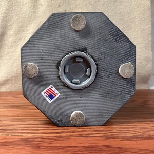 May include: A gray, octagonal wooden object with a central plastic fitting and four felt pads. A small sticker reading "MADE IN U.S.A." is affixed to the side. The object is displayed on a wooden surface.