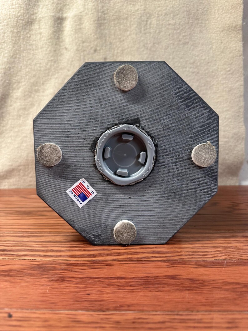 May include: A gray, octagonal wooden object with a central plastic fitting and four felt pads. A small sticker reading "MADE IN U.S.A." is affixed to the side. The object is displayed on a wooden surface.