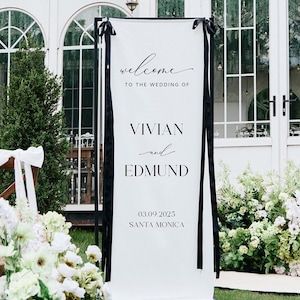 May include: A white wedding welcome sign with black detailing, displaying the names Vivian and Edmund, and the date 03.09.2023, Santa Monica. The sign is framed with black ribbons, set on a green lawn, with floral arrangements and candles.