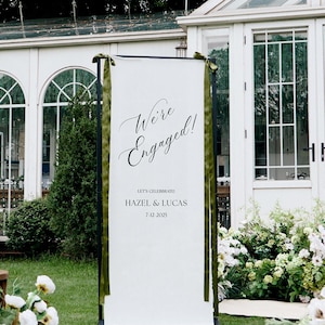 Könnte beinhalten: Ein weißes Banner mit der Aufschrift "We're Engaged!" in eleganter Schrift, mit den Namen Hazel & Lucas und dem Datum 7.4.2025. Das Banner ist mit grünen Bändern umrahmt, vor dem Hintergrund eines weißen Gebäudes und üppigem Grün.