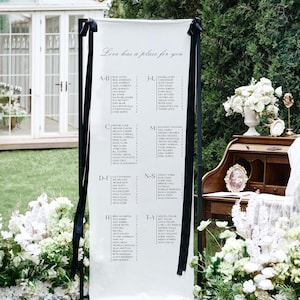 May include: A white wedding seating chart with black trim and black ribbons. The chart displays names organized alphabetically. The text "Love has a place for you" is at the top. The chart is outdoors, surrounded by greenery and white flowers.
