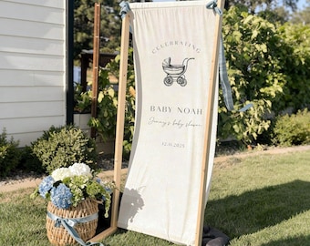 May include: A wooden framed sign with a cream-colored banner. The banner reads "Celebrating Baby Noah" with a baby carriage illustration. A wicker basket with blue and white flowers and a blue ribbon sits nearby. The sign is on a grassy lawn.