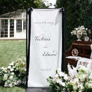 K&ouml;nnte beinhalten: Ein wei&szlig;es Hochzeits-Willkommensschild mit schwarzen R&auml;ndern und Schleifen. Auf dem Schild steht "welcome to the wedding of Victoria and Edward" mit dem Datum 2025.09.08. Wei&szlig;e Blumen und Gr&uuml;n umgeben das Schild.