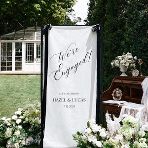 May include: A white engagement announcement banner with black trim, displaying the words "We're Engaged!" in cursive script. The names "Hazel & Lucas" and the date "7-12-2025" are also featured. The sign is decorated with white flowers.