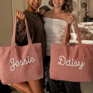 May include: Two pink corduroy tote bags with white cursive lettering. One bag says "Jessie" and the other says "Daisy". The bags have long handles and are held by two people.