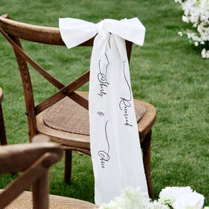 May include: Wooden chairs with woven seats are decorated with white fabric bows and personalized banners that read "Reserved" and names. The setting is outdoors on a green lawn, suggesting a wedding or special event.